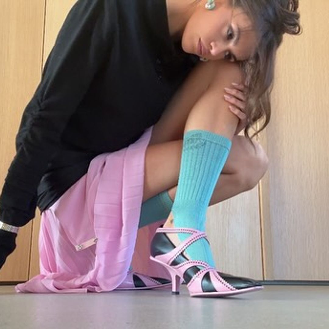 Kaia Gerber crouches in front of the camera wearing a black long sleeved top, pink skirt, black and pink pointy shoes and turquoise leg warmers. 