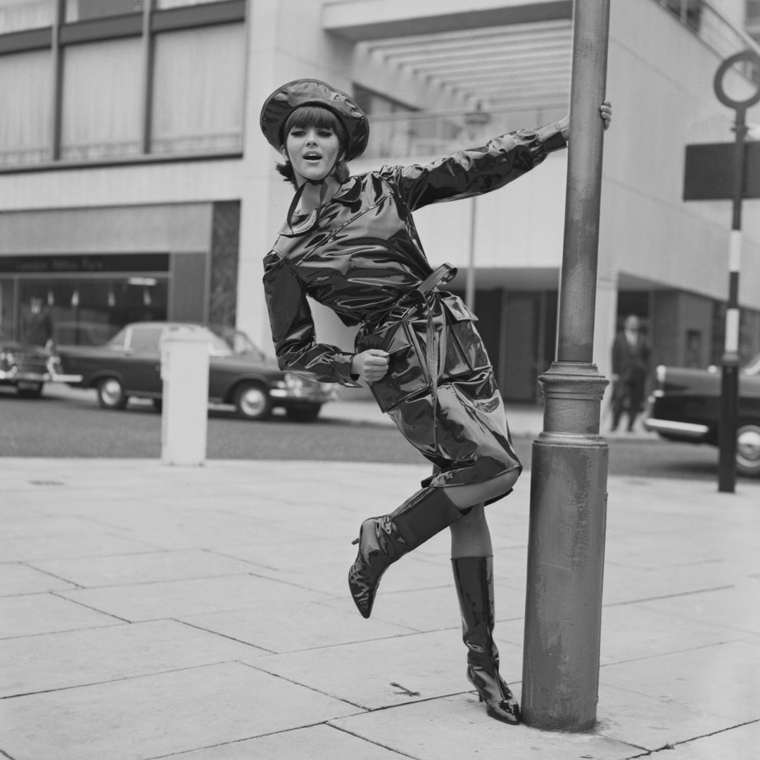 Model Jackie Bowyer swings on a lamppost wearing a black oilskin wet-weather outfit from Mary Quant, of matching coat and hat with shiny, high-heeled boots, London, 15th October 1963. The outfit was shown that evening at the first Sunday Times Internation