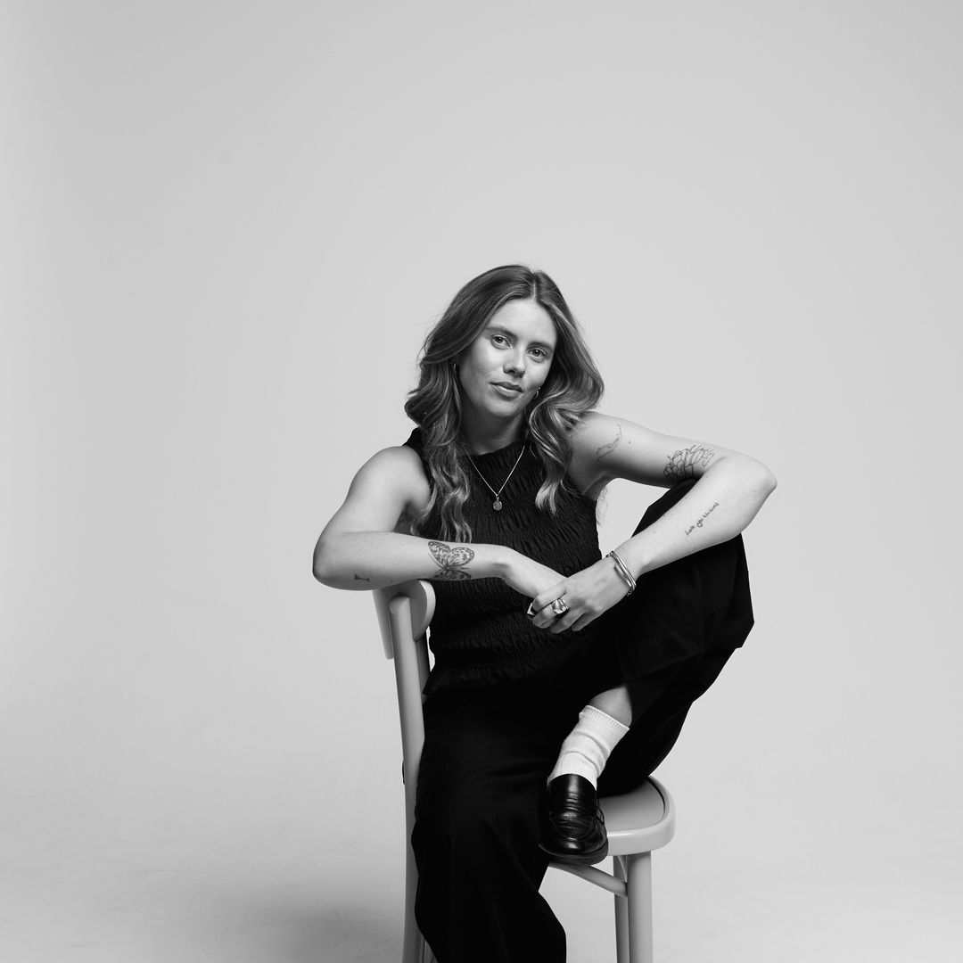 Portrait en noir et blanc d'une femme tatouée, assise nonchalamment sur une chaise, une jambe relevée, vêtue d'un haut sans manches et d'un pantalon large.