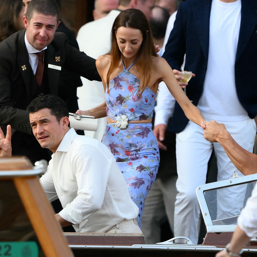 Orlando Bloom clignote un panneau de paix tout en portant une chemise blanche, rejointe par un invité dans une tenue de Halterneck florale en deux pièces, arrivant en bateau à Venise.