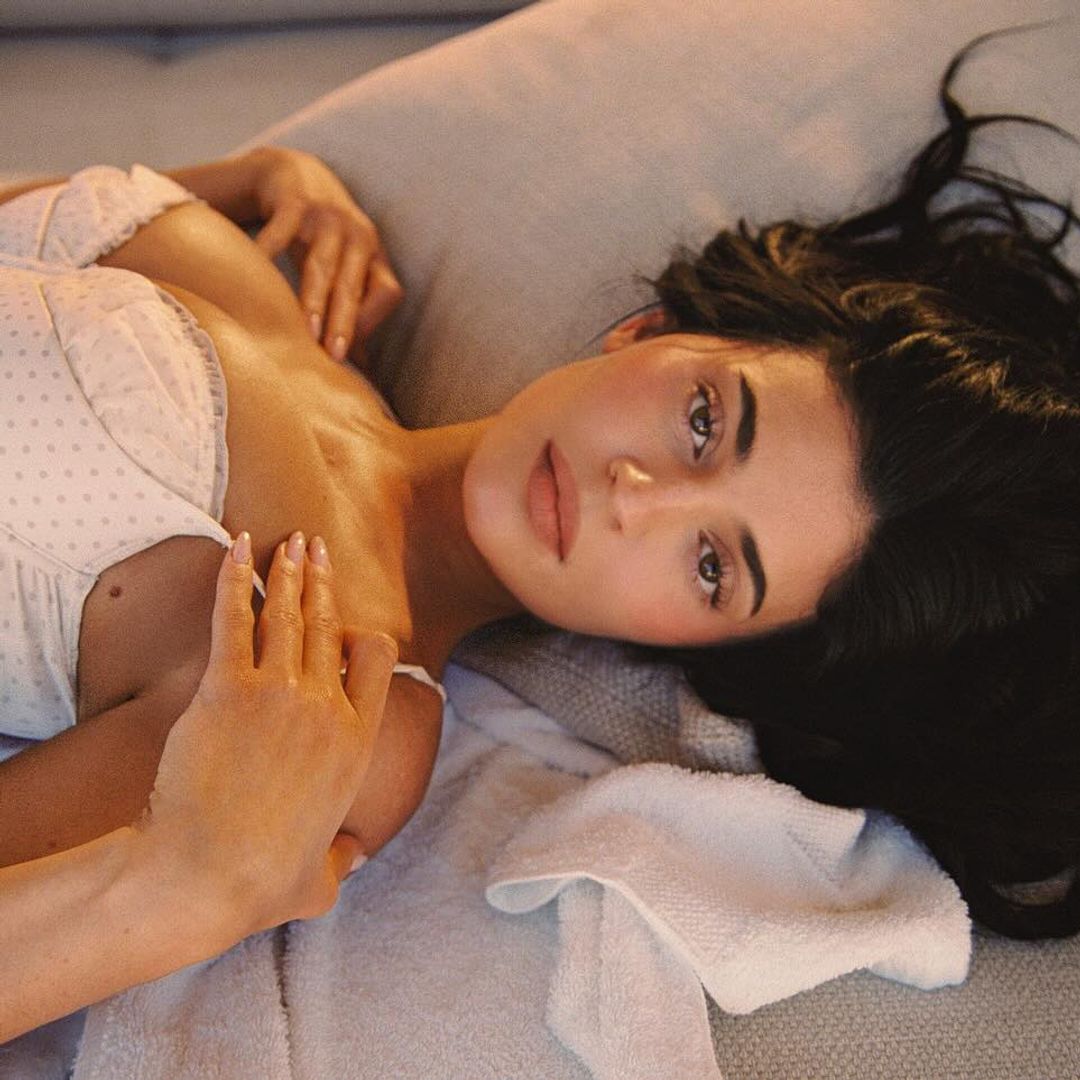 Kylie Jenner is lying on her back on a plush beige sofa, gazing directly into the camera with a serene, sultry expression. She wears a delicate white bikini that mimics vintage lingerie, featuring subtle polka-dot texture and underwire detailing reminiscent of retro brassieres. Her tousled dark hair cascades across the cushions, and her hands gently touch her chest and shoulder. Soft lighting creates a golden, intimate atmosphere, giving the scene a cinematic warmth and softness.