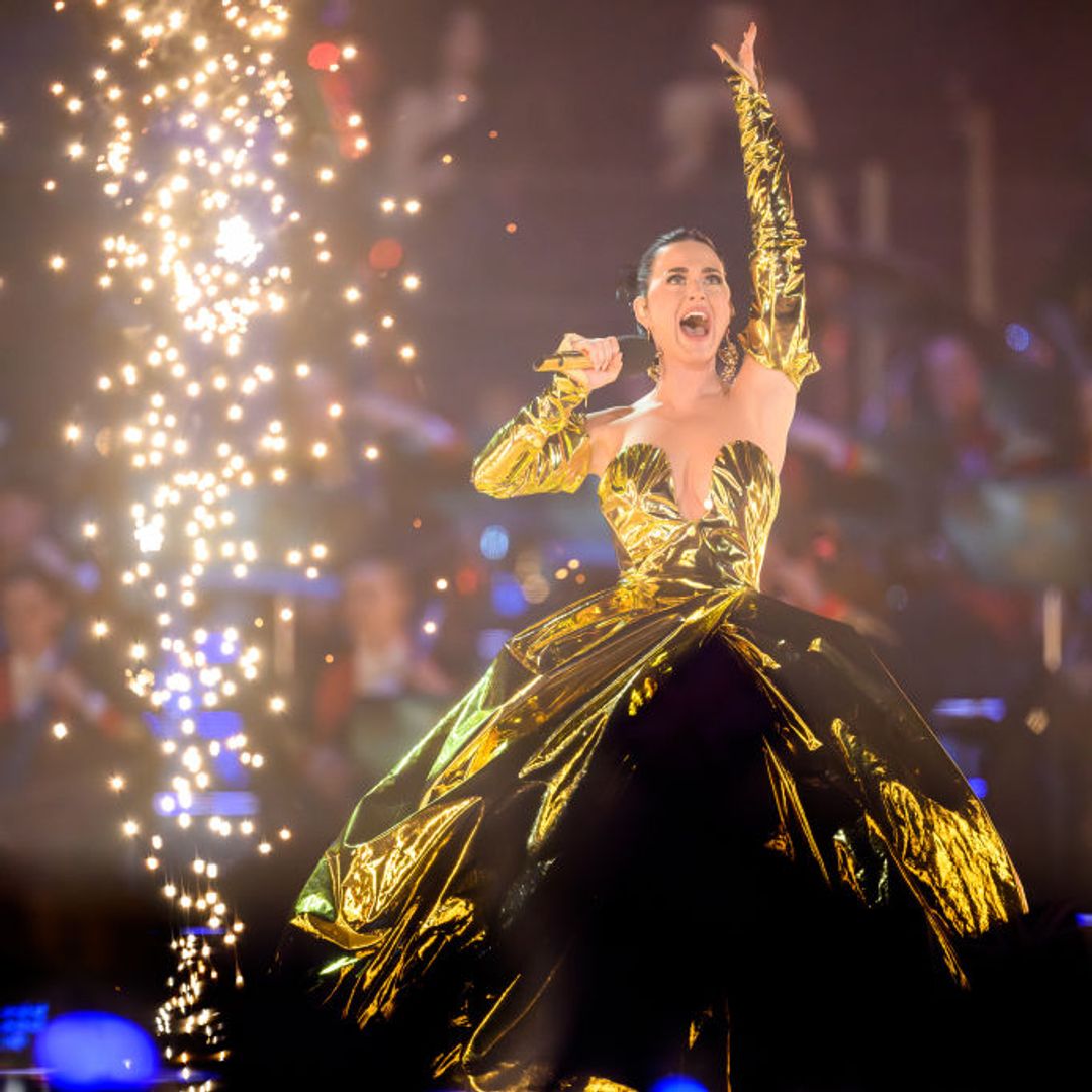 Katy Perry singing on stage in a large gold dress, hand in the air and a firework behind her
