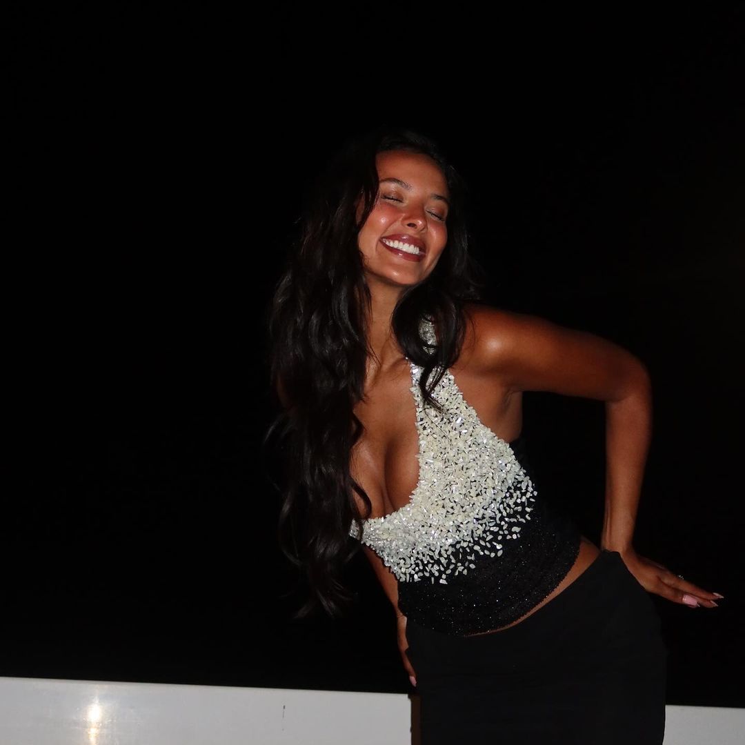 Maya Jama grins at the camera on her hotel balcony wearing a beaded white and black top and black micro mini skirt