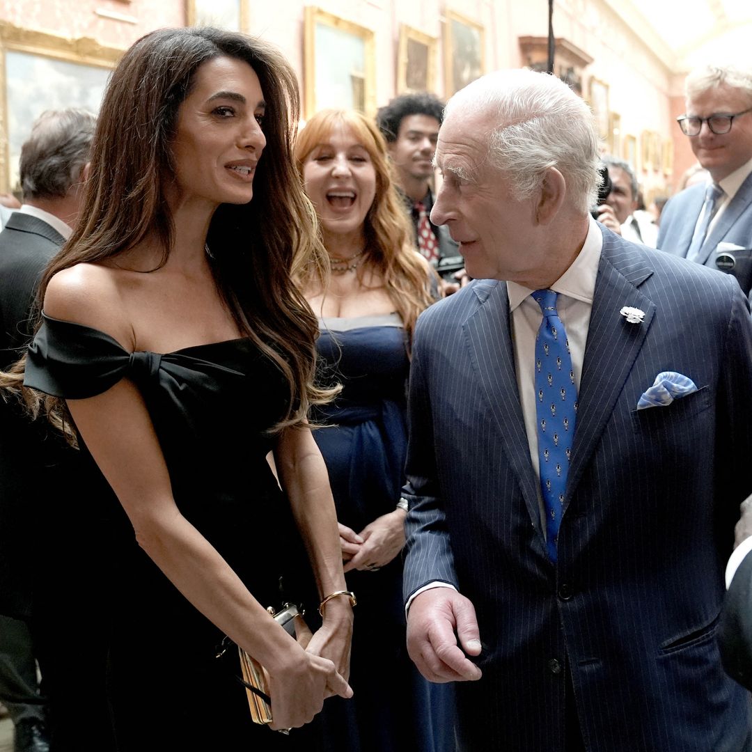 Amal Clooney and King Charles chat at Buckingham Palace. Amal wears a black off the shoulder midi dress and King Charles wears a blue pinstripe suit with bright blue tie. 