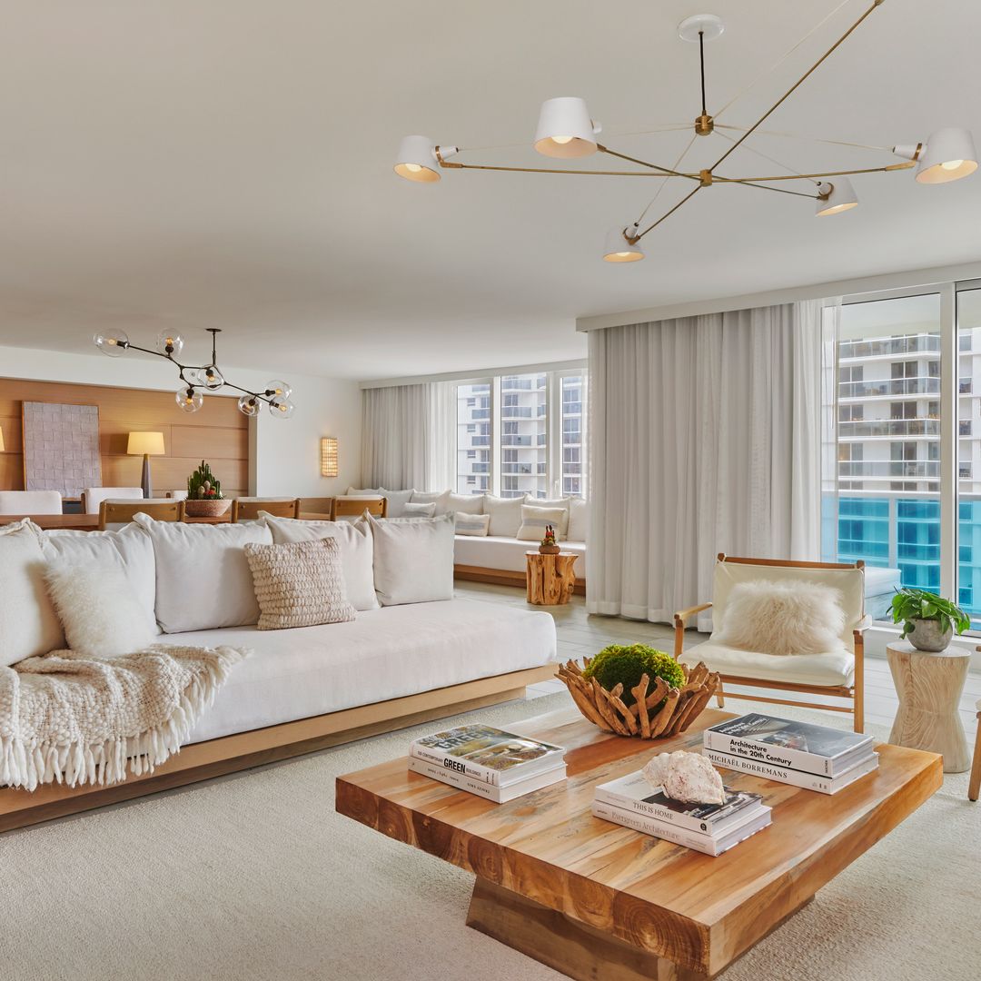 A spacious, luxurious suite with a light and airy aesthetic. The room features a large wooden coffee table surrounded by white cushioned sofas and accent chairs.