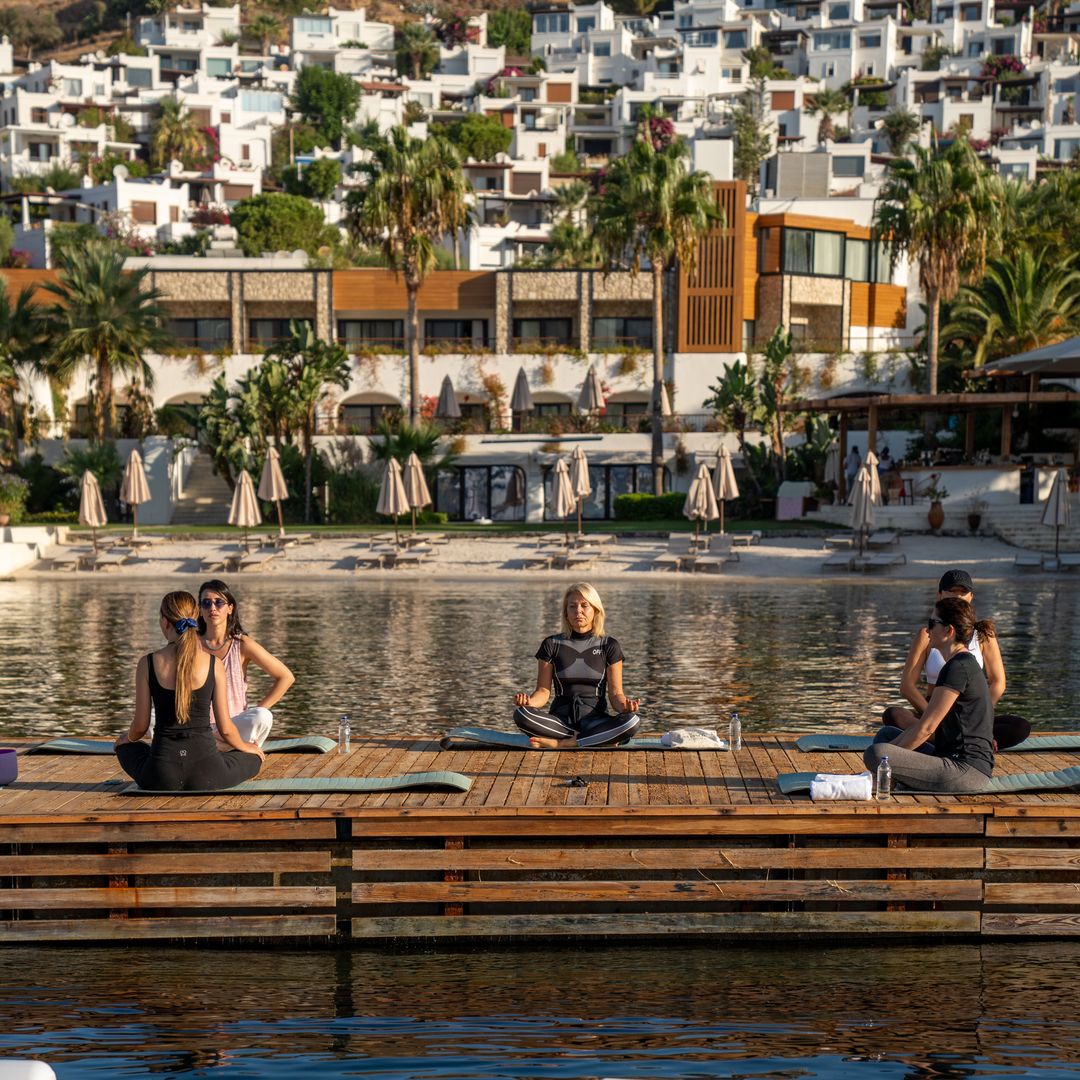Invités participant à un cours de yoga matinal sur une terrasse avec vue sur la mer à l'Avantgarde Refined Bodrum à Yalıkavak, avec l'hôtel en bord de mer et les maisons à flanc de colline en arrière-plan.