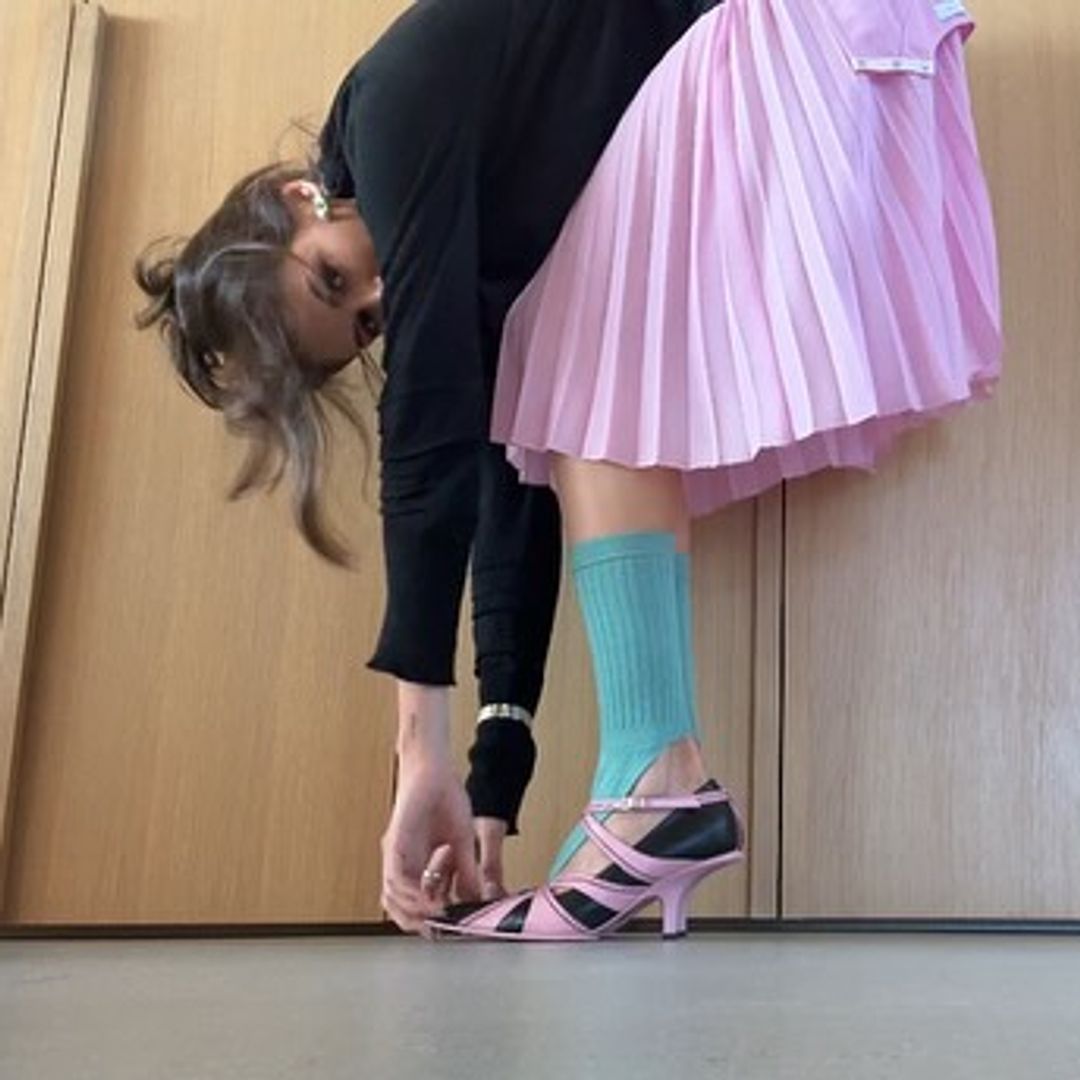 Kaia Gerber bends over and grabs her toes, looking at the camera. She wears a pink pleated skirt, black long sleeved top, black and pink pointy shoes and turquoise leg warmers