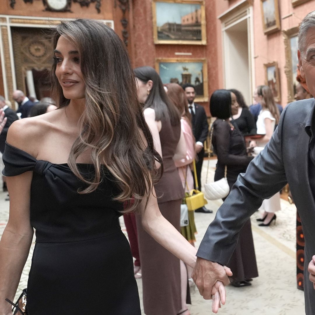 Amal Clooney and her husband George Clooney who wears a grey suit and grey open-collar shirt