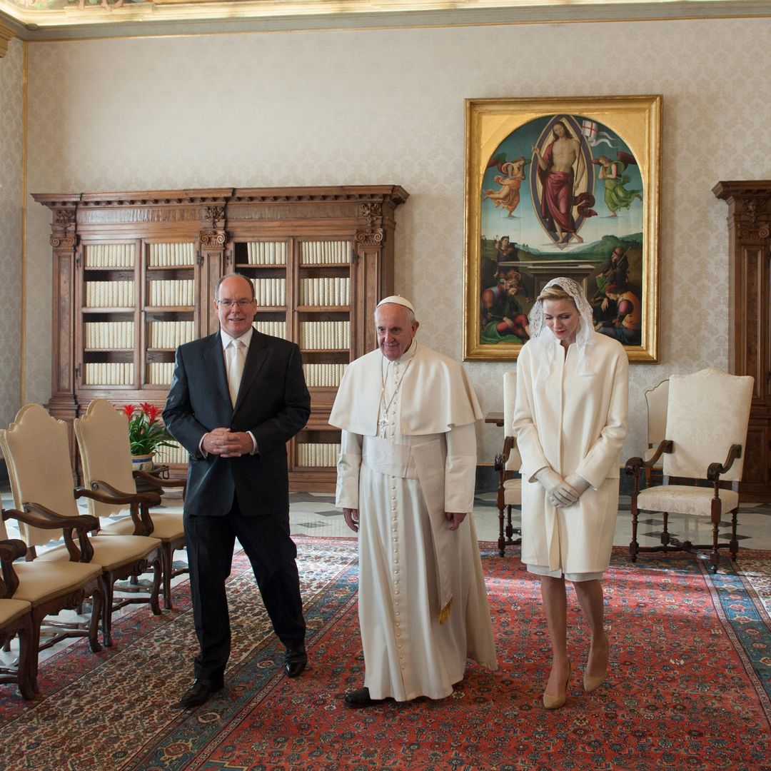 Pope Francis wears white robes and stands between Prince Albert in a black suit and Princess Charlene in a white coat and lace veil