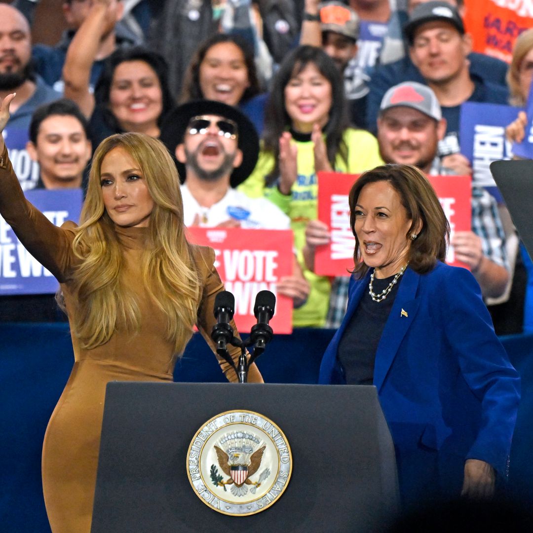 Jennifer Lopez, in a fitted brown midi dress, raises her hand alongside Vice President Kamala Harris at a rally, with a crowd holding signs reading “When We Vote, We Win.”