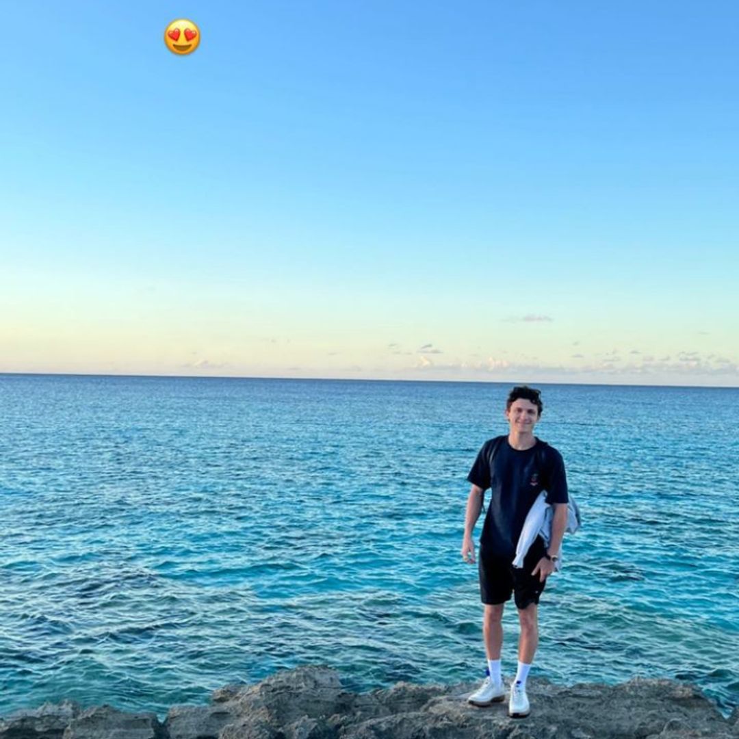 Tom Holland smiling on a cliff side with a bright blue sea and sunsetting sky behind him