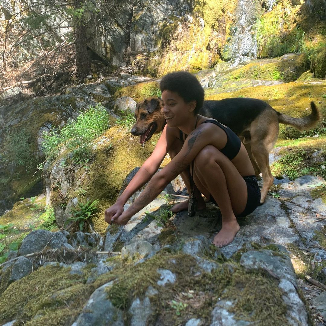 Willow Smith crouching on a mossy rock with a dog beside her