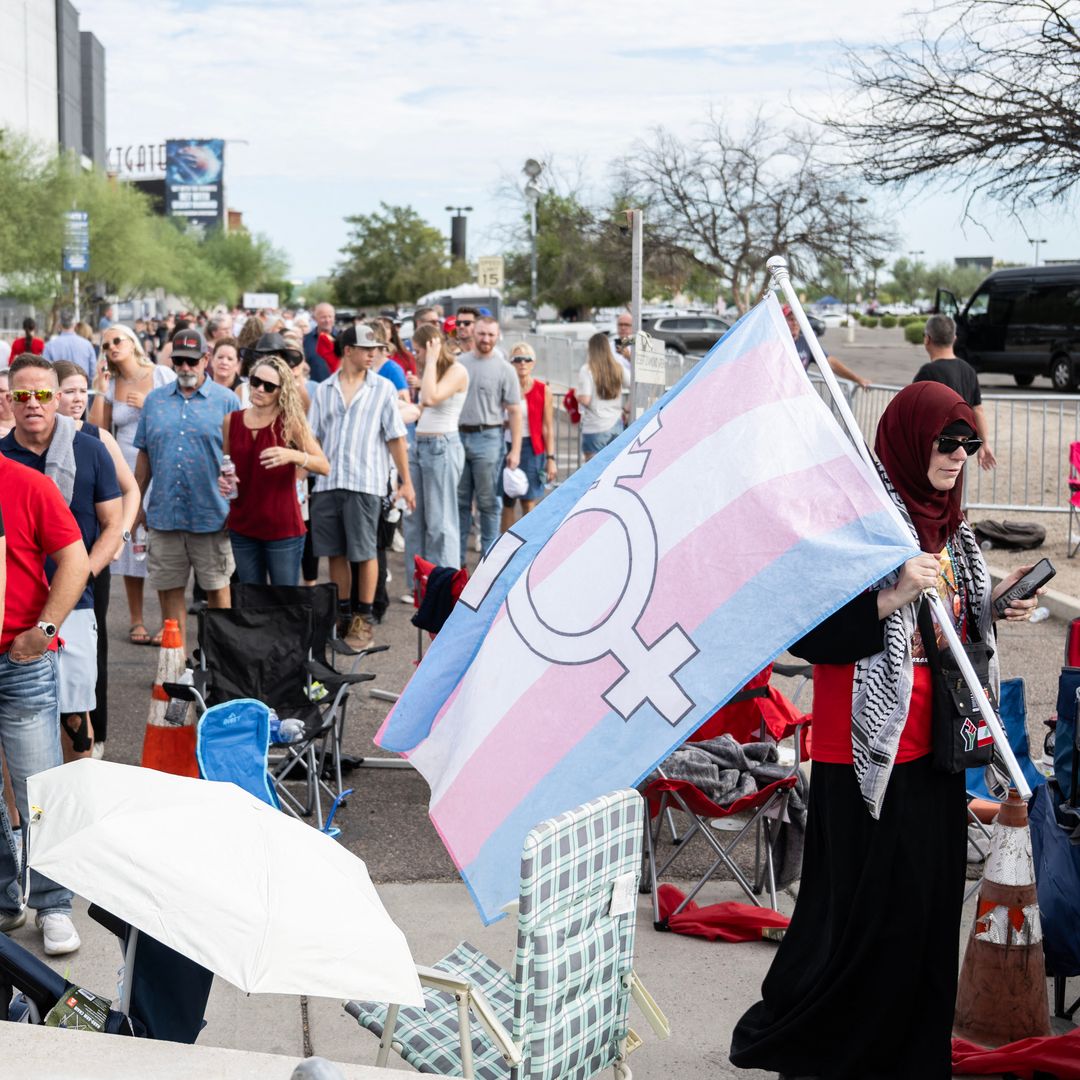A protester carrying a transgender support flag walks past people waiting to attend the memorial for Charlie Kirk at State Farm Stadium in Glendale, Arizona, on September 21, 2025.