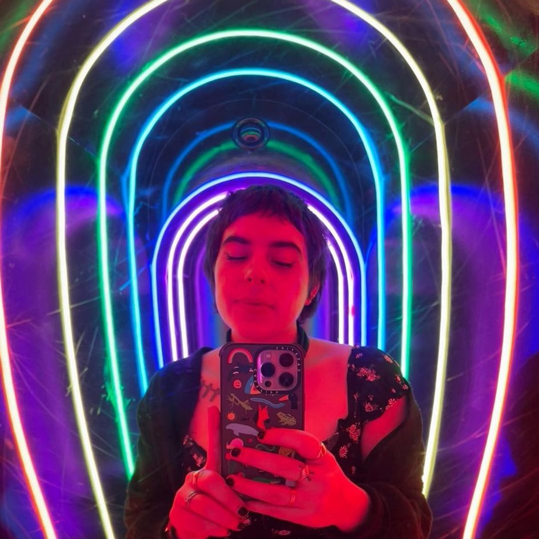 Bella posing for a mirror selfie with neon rainbow lights reflected all around her