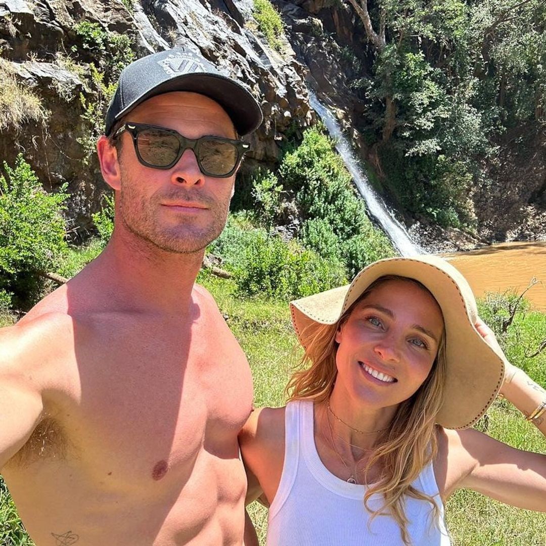 Chris and Elsa smiling for a selfie near a waterfall, both are wearing hats and her is wearing sunglasses