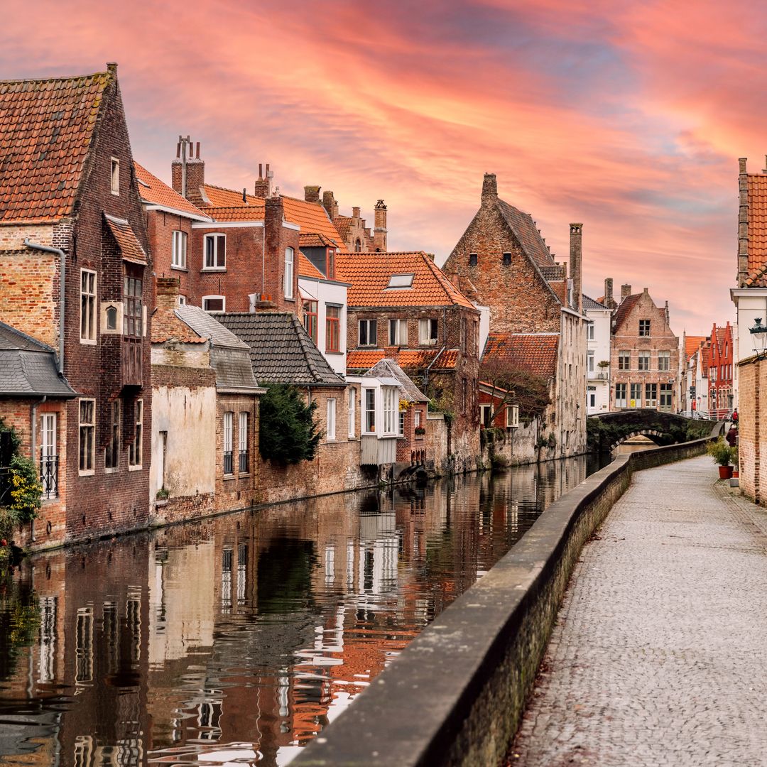 a charming European canal scene at sunset. The canal is lined with historic brick houses, some extending over the water, reflecting the warm hues of the sky.