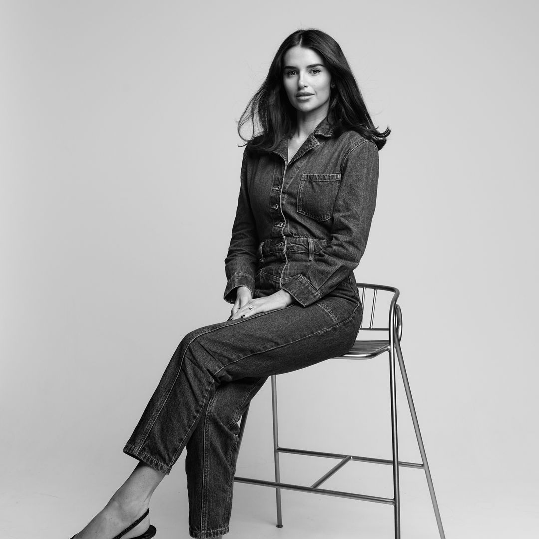 Portrait en noir et blanc d'une femme portant une combinaison en jean et des talons à bride arrière, assise sur un grand tabouret en métal sur un fond de studio uni.
