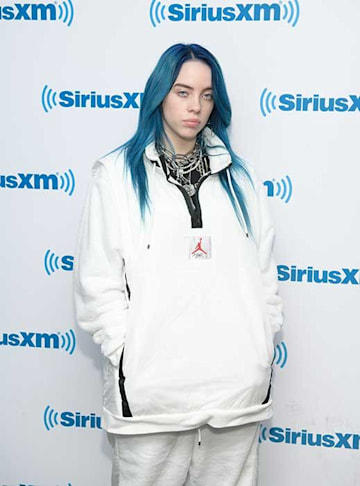 billie poses at a press event wearing an all white loose fitting tracksuit which stands out against her blue hair and she looks relaxed 