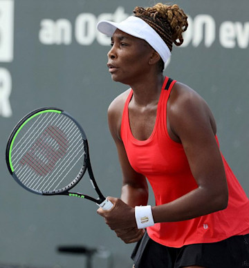 venus is holding a tennis racket and focused on something in front of her as she wears a red vest top that shows off her tined physique and a white visor shields her eyes from the sun