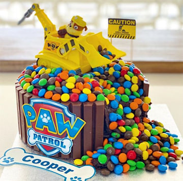 A chocolate cake with colorful round sweets poured on top and a yellow excavator toy spilling over it stands on a table