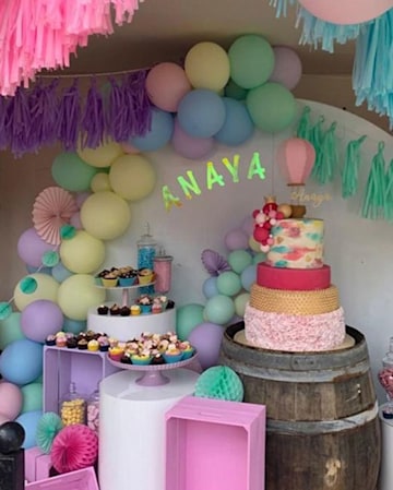 A large four-tiered cake in pink and gold tones rests on a wooden barrel, surrounded by balloon arches and tissue paper streamers, with cup cakes nearby.