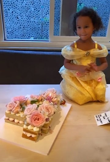 A little girl kneeling on the floor wearing a yellow dress that looks like the dress of the character Belle from the Disney movie Beauty and the Best, with the floor beside her filled with cream and topped with pink flowers I have a cute sponge cake.