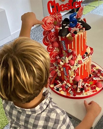 Photo of a tall red and white striped cake with a blond haired boy reaching for it with his back to the camera