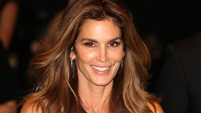headshot of cindy crawford smiling at the camera as her long brown hair blows back slightly from her face which boasts a natural makeup look with discreet smoky effect eye makeup