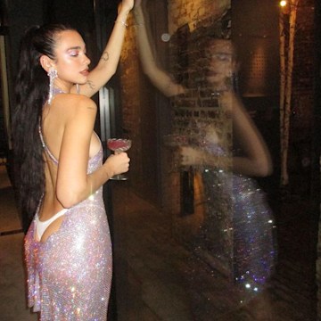 Dua Lipa holds a coupe of alcohol and poses in a backless thong-baring dress