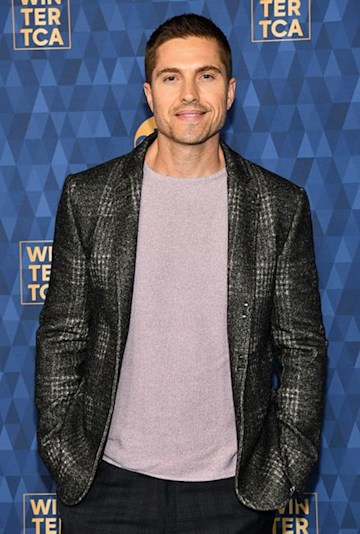 Eric Winter wearing a grey blazer and a pale lilac top at a press event. 