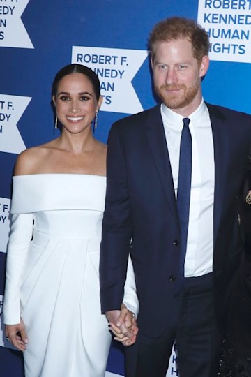 Meghan in a white off-the-shoulder dress with her hair slicked back holding hands with Harry as they smile at the camera in front of a blue and white screen