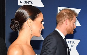 Meghans head and shoulders in profile showing a braided bun with shiny slicked back hair and sparkly earrings with Prince Harry in the background also in profile