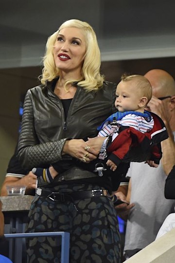 gwen stands in an auditorium holding her baby boy on her hip who is wearing a stripy top and she wears all black with a touch of dark red lipstick