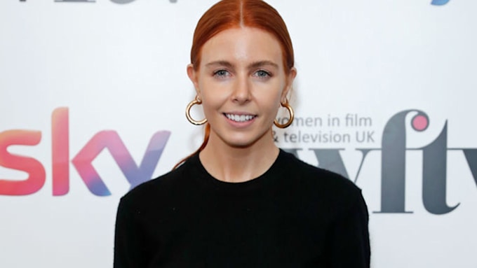 Stacey Dooley wearing hoop earrings and a black crew neck jumper, she's smiling. 