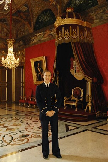 a vintage colour photo of a man dressed in a black and gold buttoned uniform in a marble floored room that has burgundy walls and a red and gold throne stands in the background