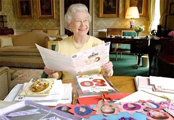 the queen sits at a large round wooden table positioned in a room adorned with soft furnishings and green carpet