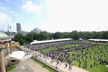 a view of buckingham palace garden with trees and the london skyline in the distance and the edge of the stone pillars of the palace just visible to the left as crowds gather on the lawn