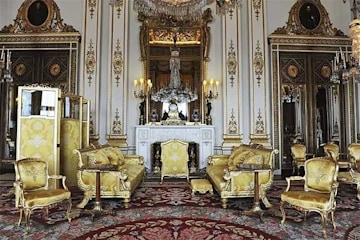 a white walled room with exceptionally high ceilings is filled with gold chairs and sofas positioned throughout the room