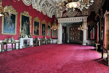 the a large room decorated with deep red carpet and matching wallpaper with small wooden tables pushed to the edges of the room below gold framed paintings