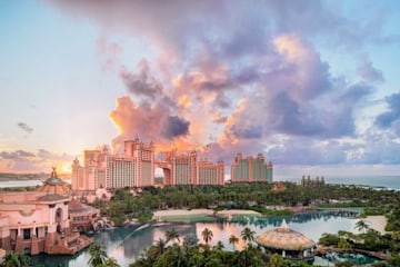 a view of a grand hotel set on the beach with a large pool and palm trees ascends into the pink and peach hued clouds at sunset